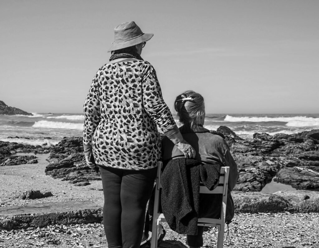 two women on a beach one sitting, one standing with her arm around the other in support of each other